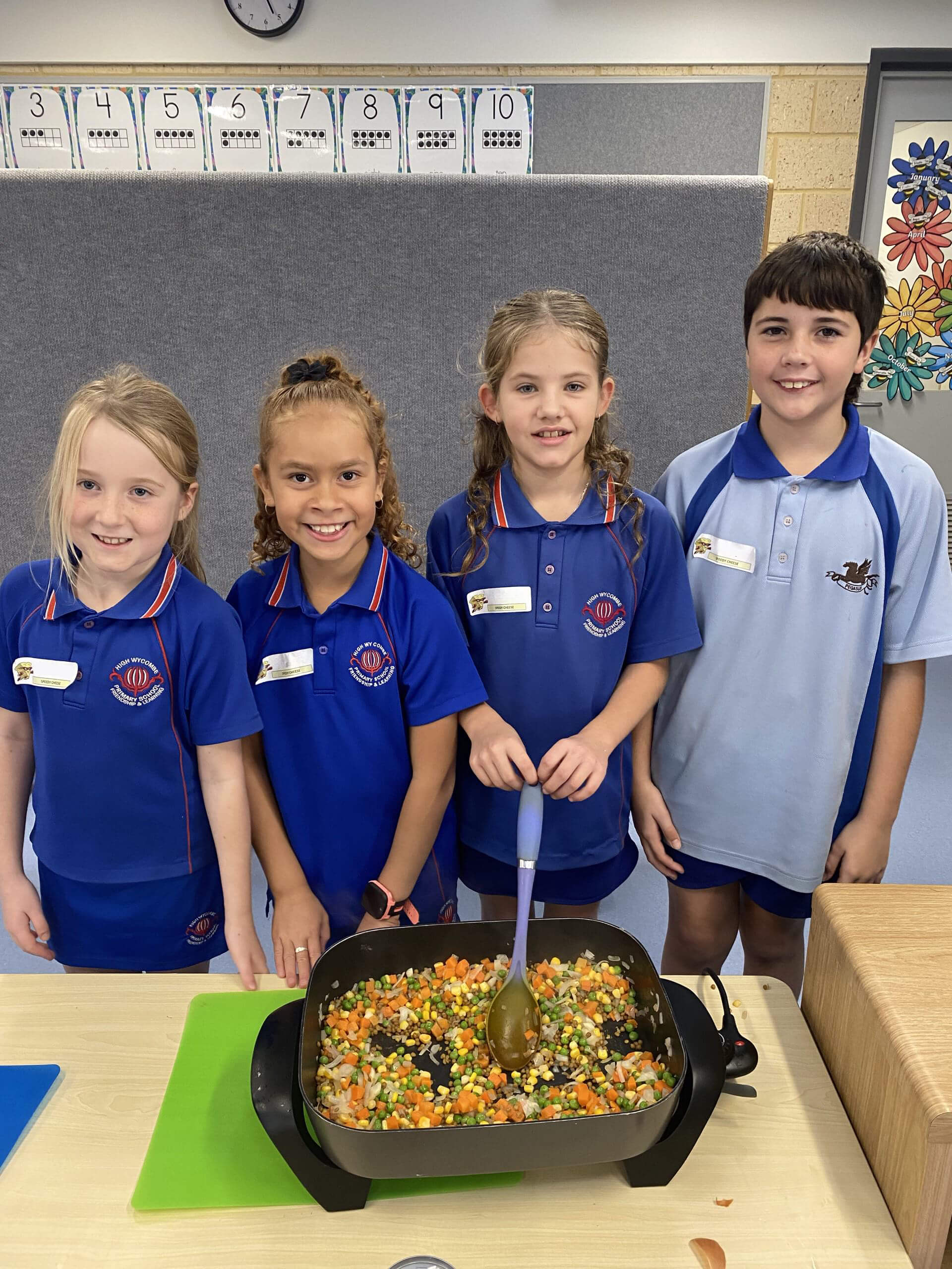 The image shows four children standing in a classroom, smiling at the camera while participating in a cooking activity. They are all wearing blue school uniforms with name tags. In front of them is an electric skillet filled with a colorful mixture of chopped vegetables, including carrots, corn, peas, and beans, being stirred with a large spoon. The children appear proud and engaged in the activity. Behind them, the classroom wall is decorated with number charts and colorful artwork, creating a bright and educational atmosphere. The overall scene is one of teamwork and learning through hands-on experience.