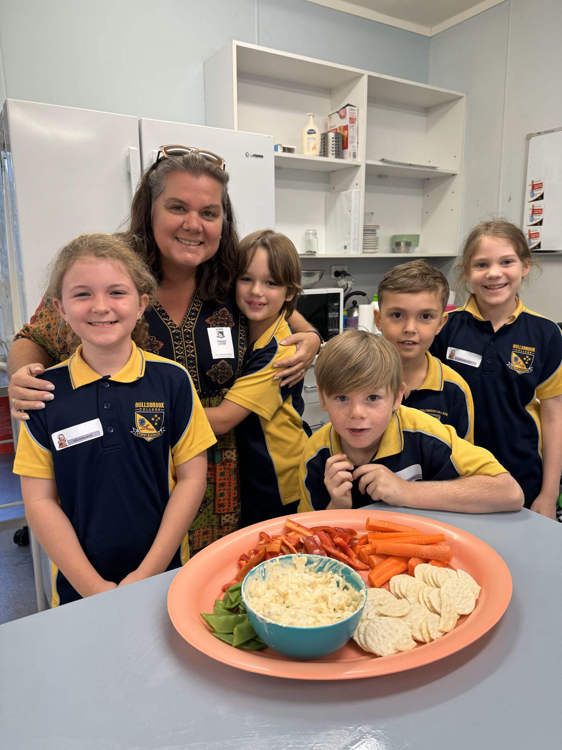 The image shows a group of children and an adult, likely a teacher or caregiver, standing together in what appears to be a kitchen or classroom setting. The children are wearing matching school uniforms, consisting of dark blue shirts with yellow collars and trim. The adult is wearing a patterned blouse and has her arms around some of the children, smiling at the camera.
In front of the group is a table with a large, round platter containing various snacks. The platter includes sliced carrots, red bell peppers, snap peas, and a bowl of dip, along with some rice crackers. The setting is bright and cheerful, with kitchen supplies visible on the shelves in the background. The overall mood of the image is positive and engaging, suggesting a communal activity such as a cooking class or snack time.