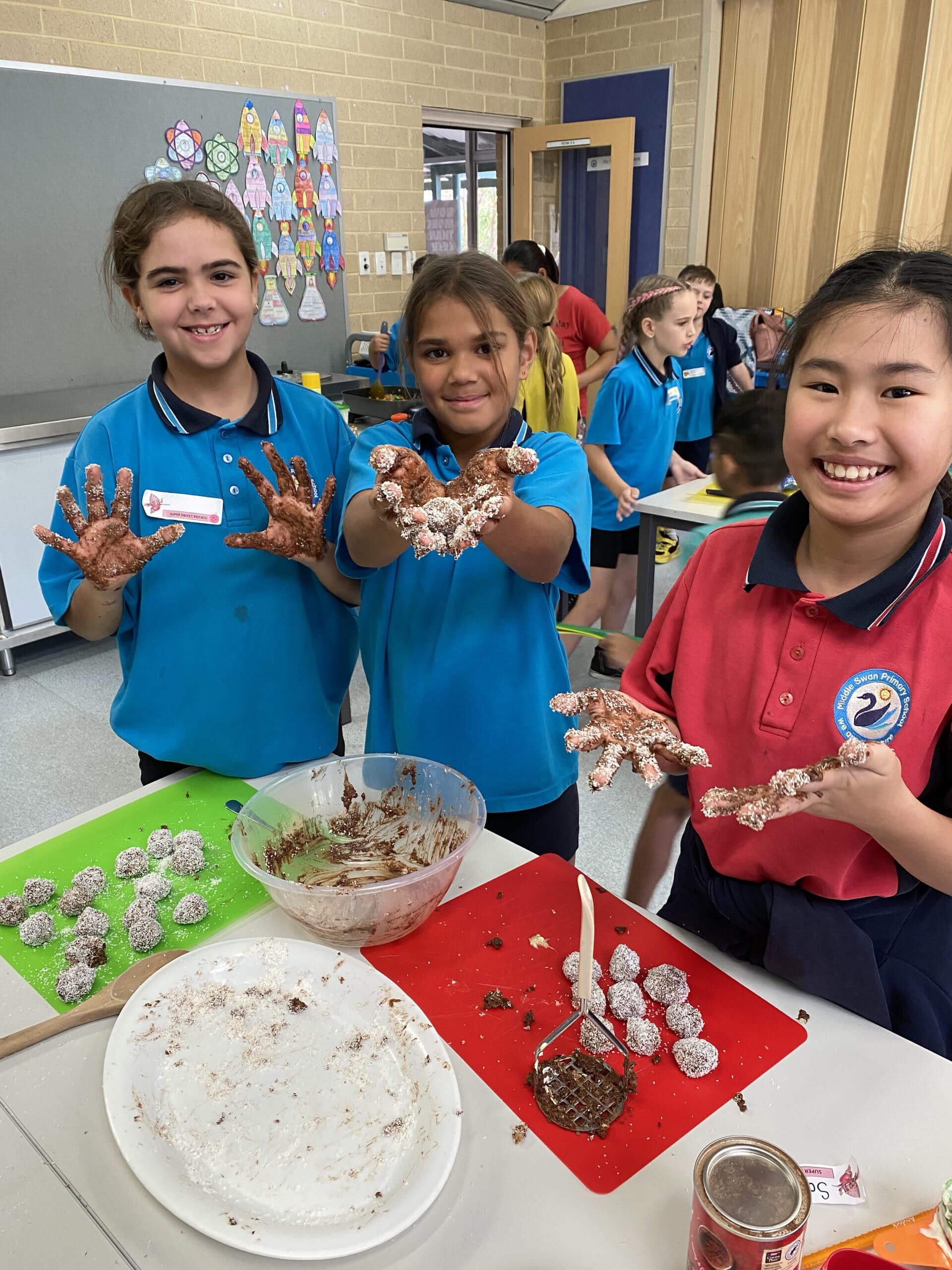The image shows three girls in a classroom setting, enthusiastically participating in a cooking activity. They are smiling and showing their hands covered in what looks like chocolate and powdered sugar. The girls are wearing school uniforms with name tags; two of them are in blue shirts, while the third is in a red shirt. In front of them on the table are mixing bowls, a wooden spoon, a masher, and plates filled with round chocolate-coated treats they have made. Other children are visible in the background, working on similar activities, creating a lively and engaging atmosphere. The classroom walls are decorated with colorful artwork, adding to the cheerful environment.