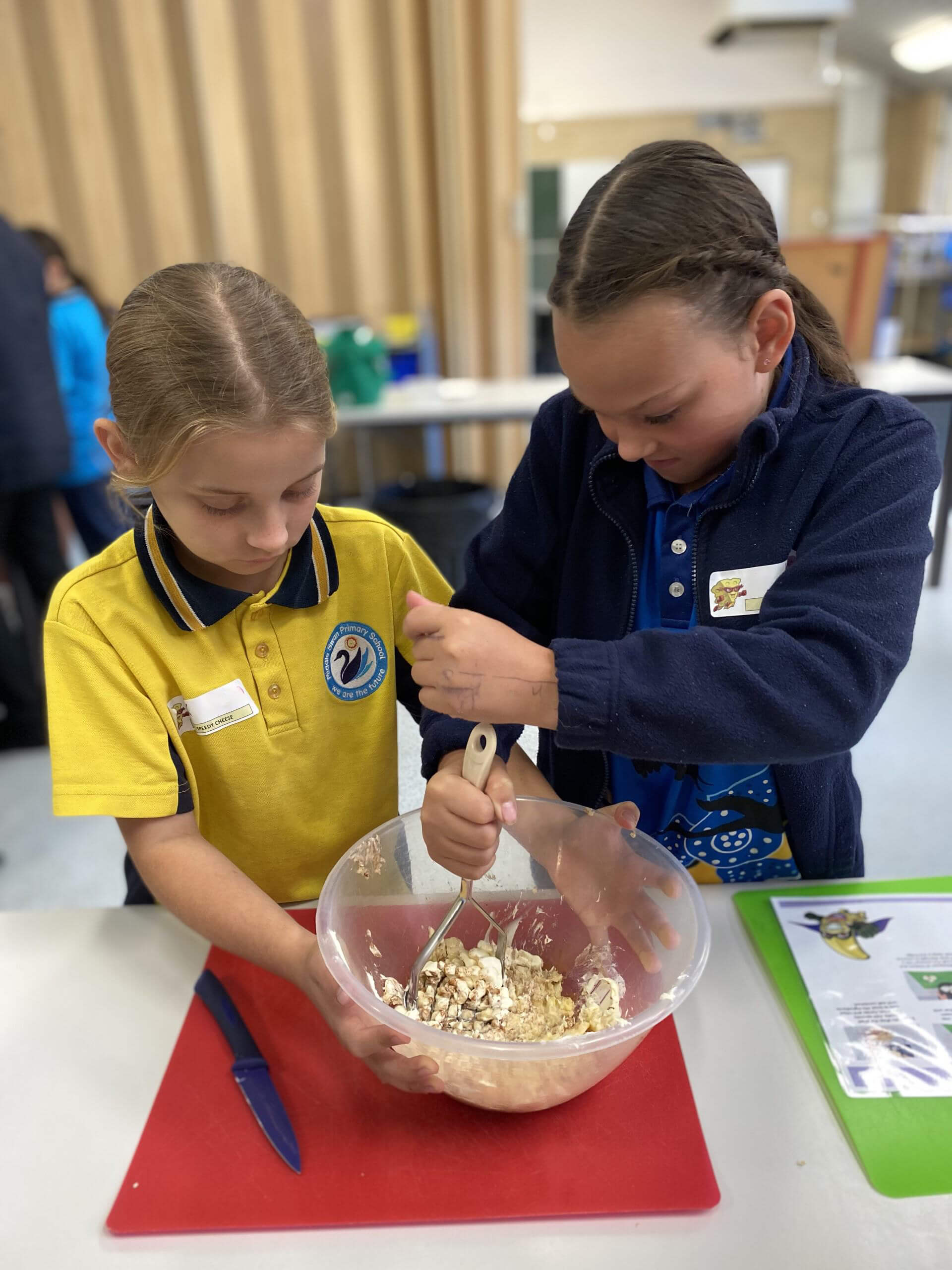 The image shows two young girls engaged in a cooking activity. They are standing at a table and working together to mix ingredients in a large, clear plastic bowl. One girl is holding the bowl steady, while the other is using a masher to combine the ingredients.
The girl on the left is wearing a yellow school polo shirt with a school emblem, and she has her hair tied back. The girl on the right is wearing a dark blue jacket over a blue shirt and is also focused on mixing the ingredients.
On the table in front of them, there is a red cutting board with a blue knife, and next to it is a green placemat with what appears to be a recipe or instructions sheet. The background shows a classroom or kitchen setting, with other students and tables visible, indicating a group activity or cooking class. The atmosphere seems educational and collaborative.