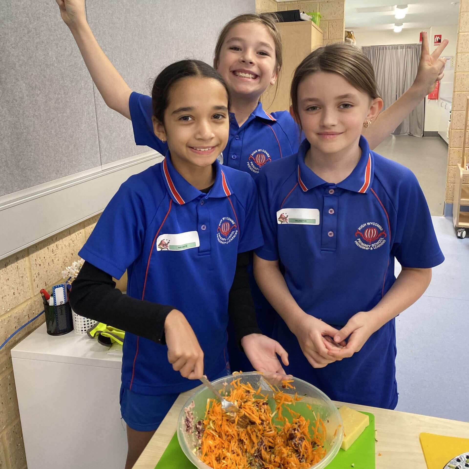 The image shows three young girls in a school setting, wearing blue school uniforms. They are grouped around a table with a large bowl of freshly made salad, which includes ingredients like shredded carrots and purple cabbage. One girl is mixing the salad while the other two are posing playfully with peace signs. The room has a modern feel with a light wood paneling and a fabric partition in the background. There’s a hint of classroom environment with a few educational items visible on shelves and a wall-mounted board.