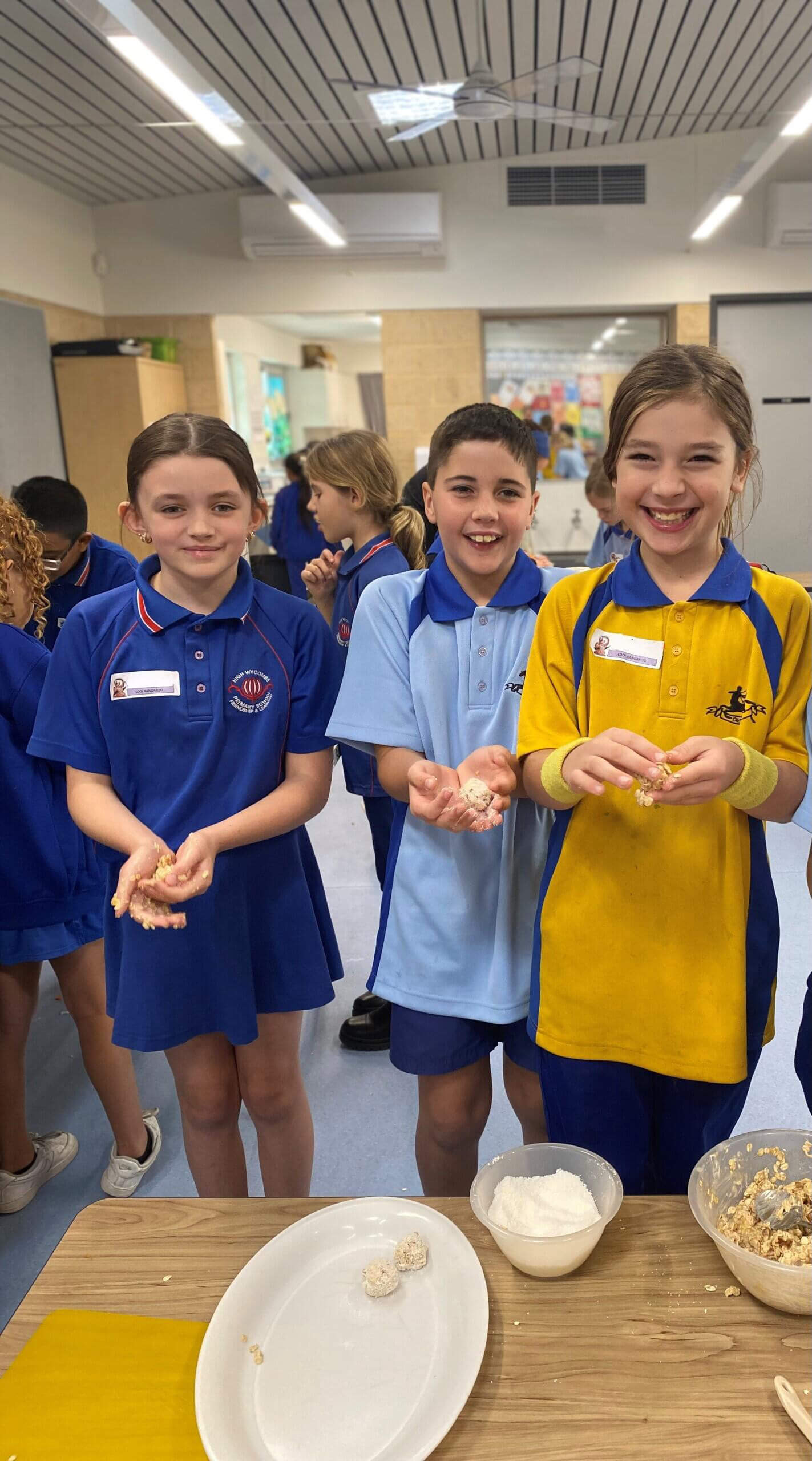 The image shows three children in a brightly lit classroom or kitchen setting, actively participating in a cooking activity. The children, two girls and a boy, are smiling and holding food mixture in their hands, proudly displaying their work. They are wearing different colored school polo shirts with name tags, indicating a collaborative school event. In front of them on a wooden table are a mixing bowl, a bowl of white powder, and a plate with a few pieces of their food creation. Other children in blue uniforms are visible in the background, further emphasizing the group activity and cheerful atmosphere.